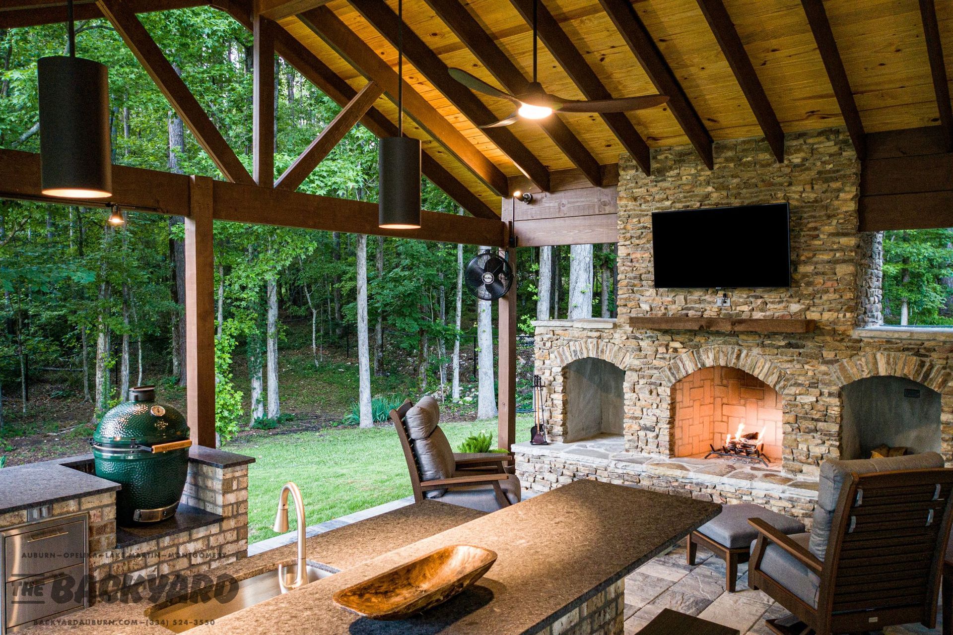 Outdoor living area with stone fireplace, bar, Big Green Egg grill, and TV overlooking a lush green lawn.