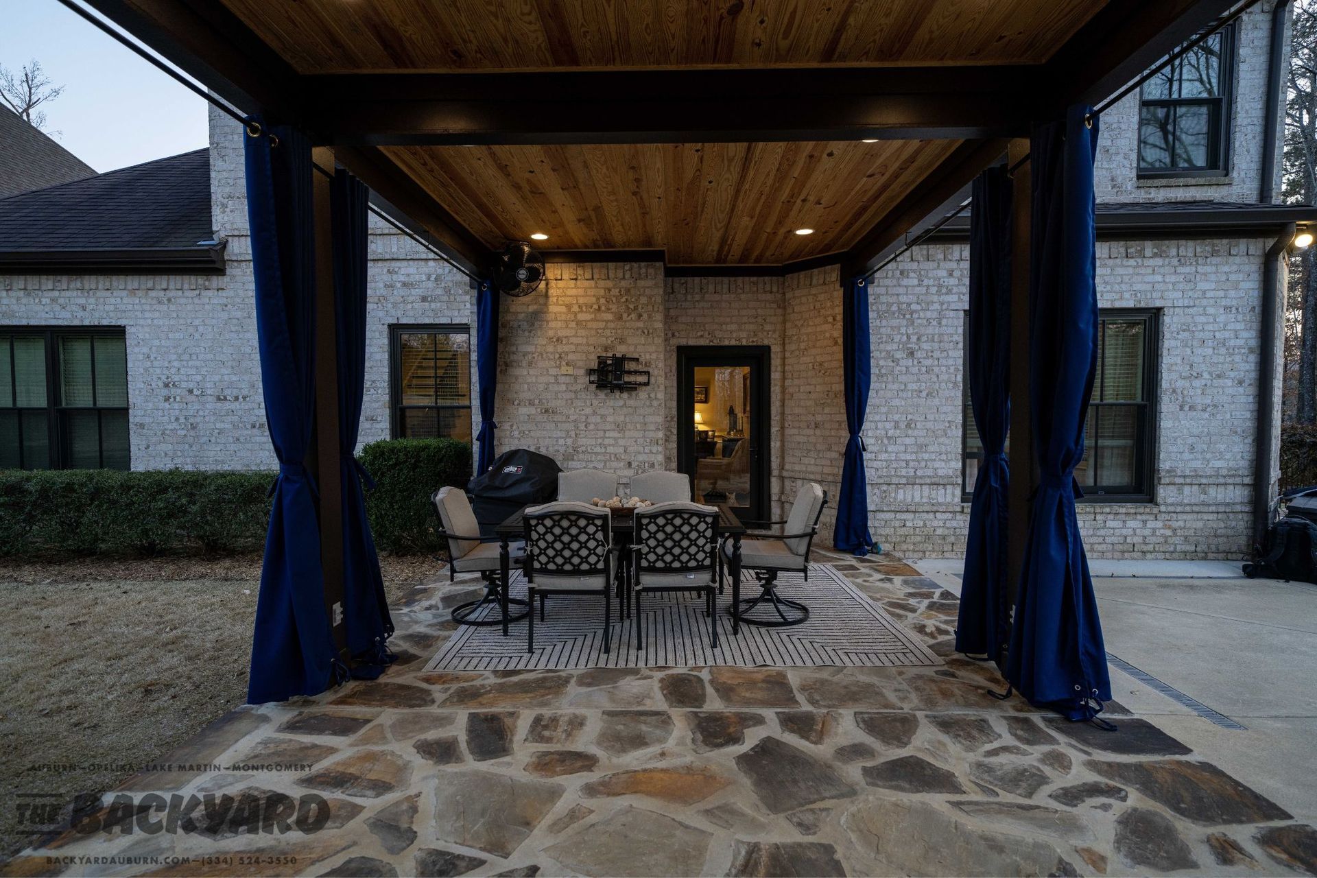 A patio with a dining set under a wooden pergola, blue curtains, brick house, and a stone floor.