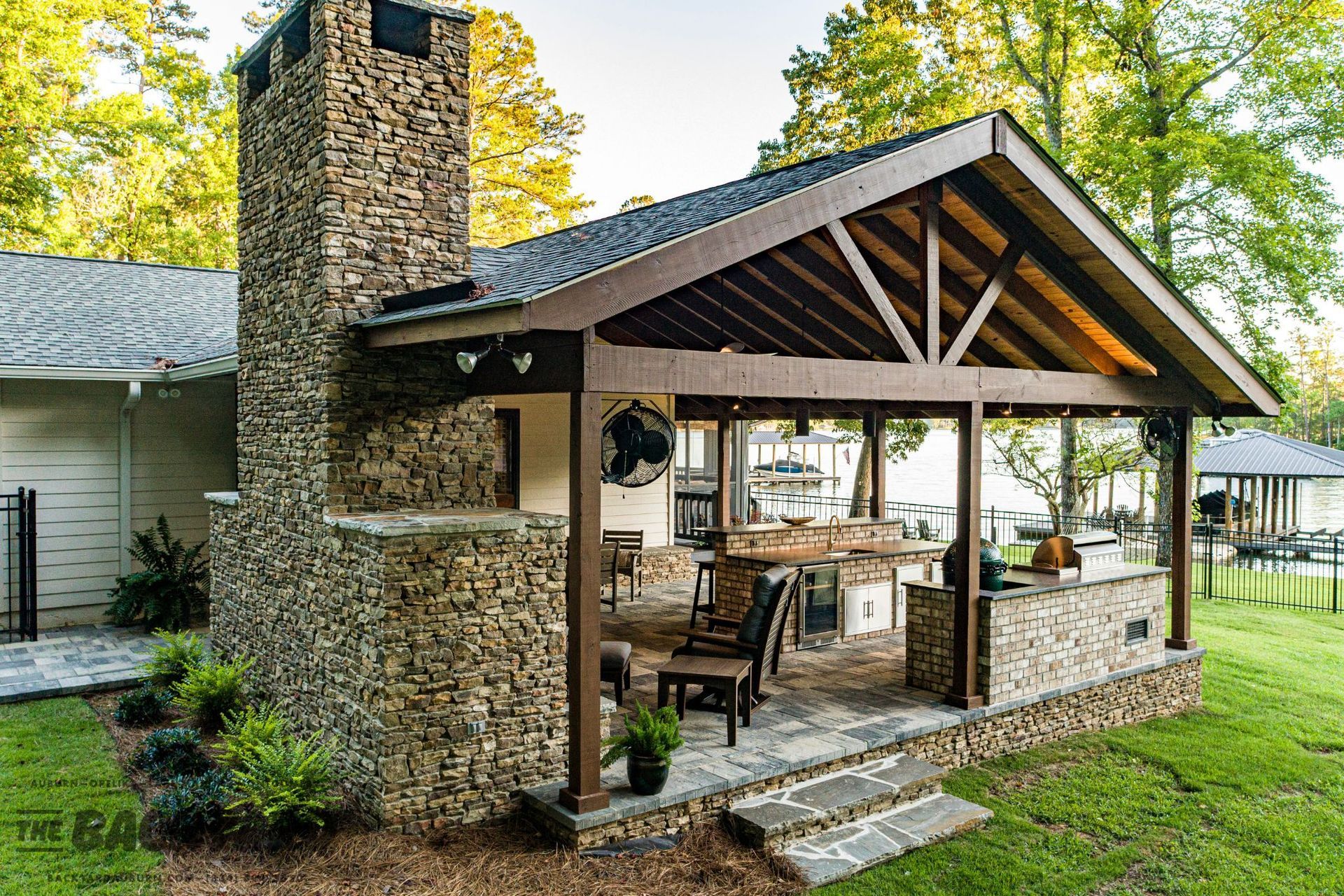 Outdoor kitchen with stone chimney, wooden beams, and lake view.
