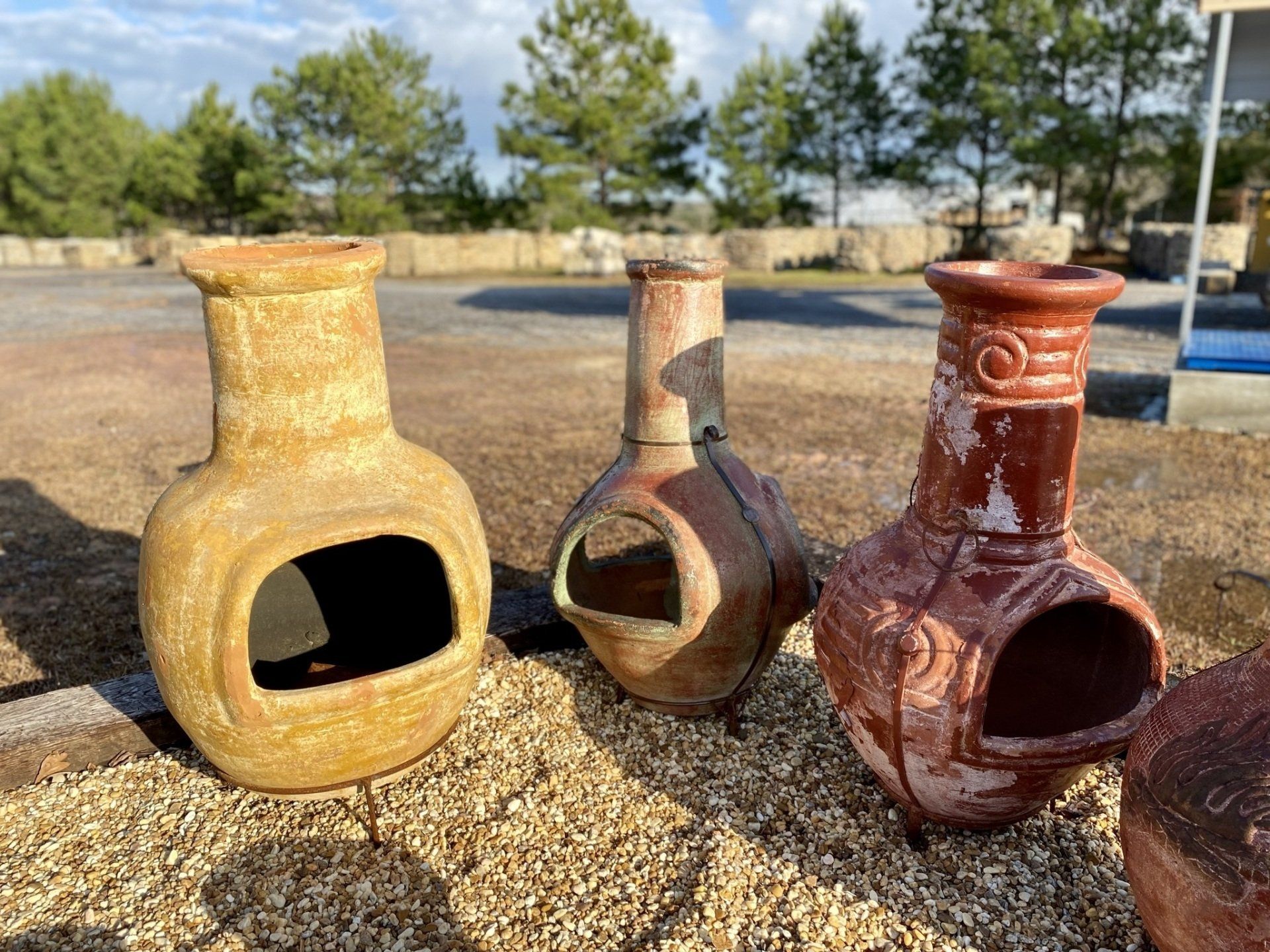 Three terracotta chimineas, various colors, on a bed of gravel, with trees and a blue sky in the background.