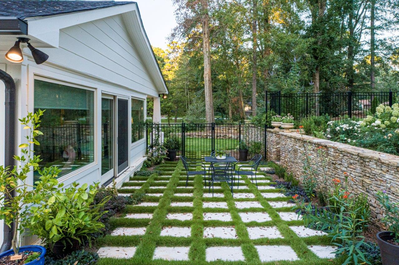 Patio with square grass tiles, table, and chairs next to a white house and stone wall with flowers.