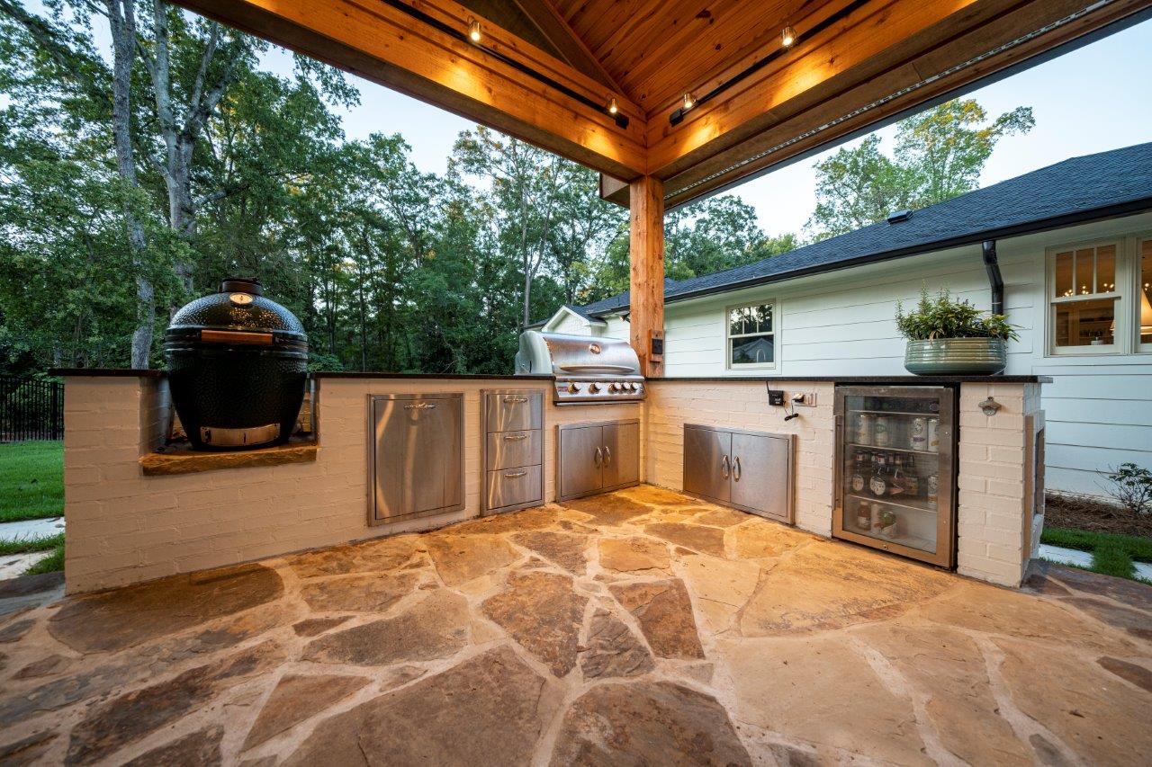 A large outdoor kitchen with a grill and a refrigerator under a covered patio.