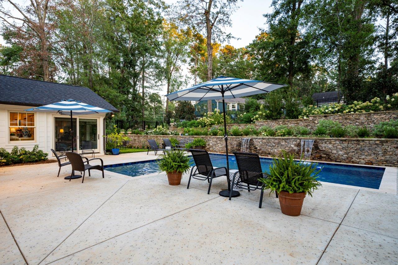 Backyard with pool, patio, and lounge chairs. Two striped umbrellas provide shade.