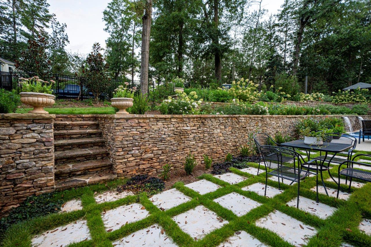 Patio with stone wall, stairs, and checkered grass design. Table and chairs. Landscaping with flowers.