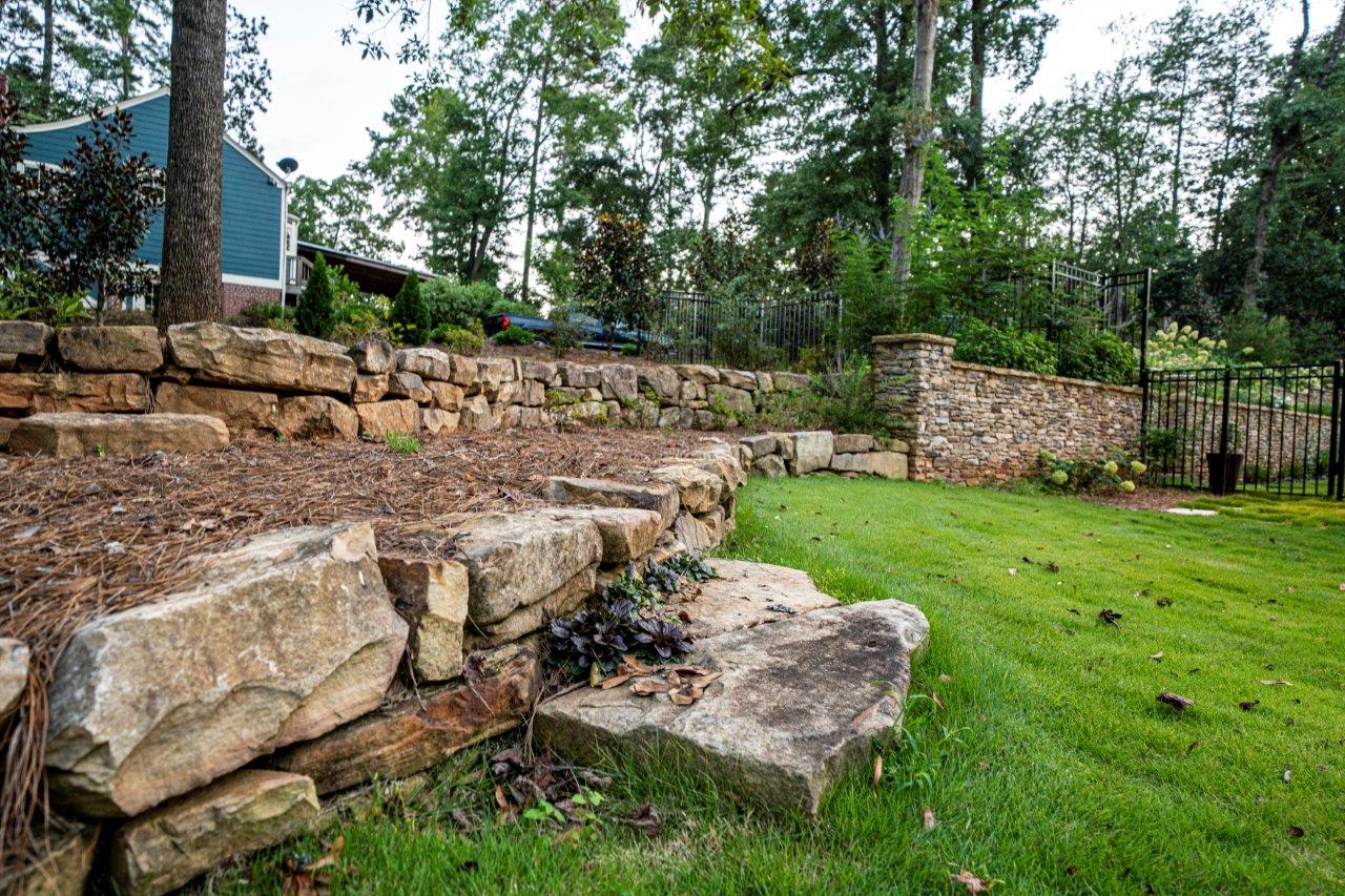 Stone retaining wall with green grass and trees in a backyard.