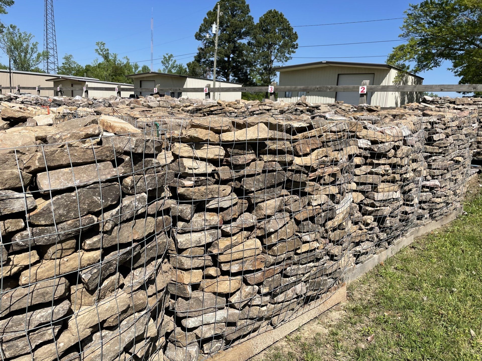 Stone blocks in a wire cage form a wall. Green grass and buildings are in the background under a sunny sky.