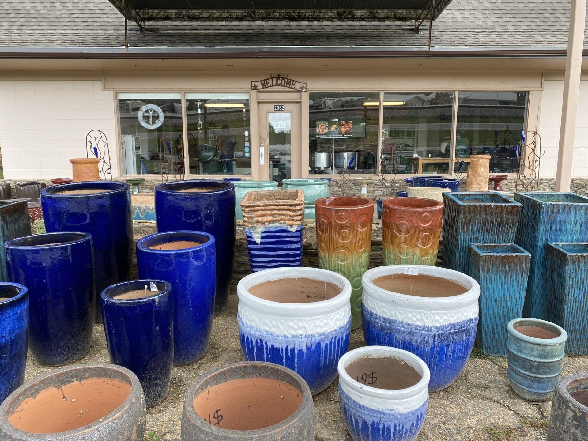 Rows of colorful ceramic pots in front of a building with windows. Variety of shapes and glazes, including blue, brown, and white.