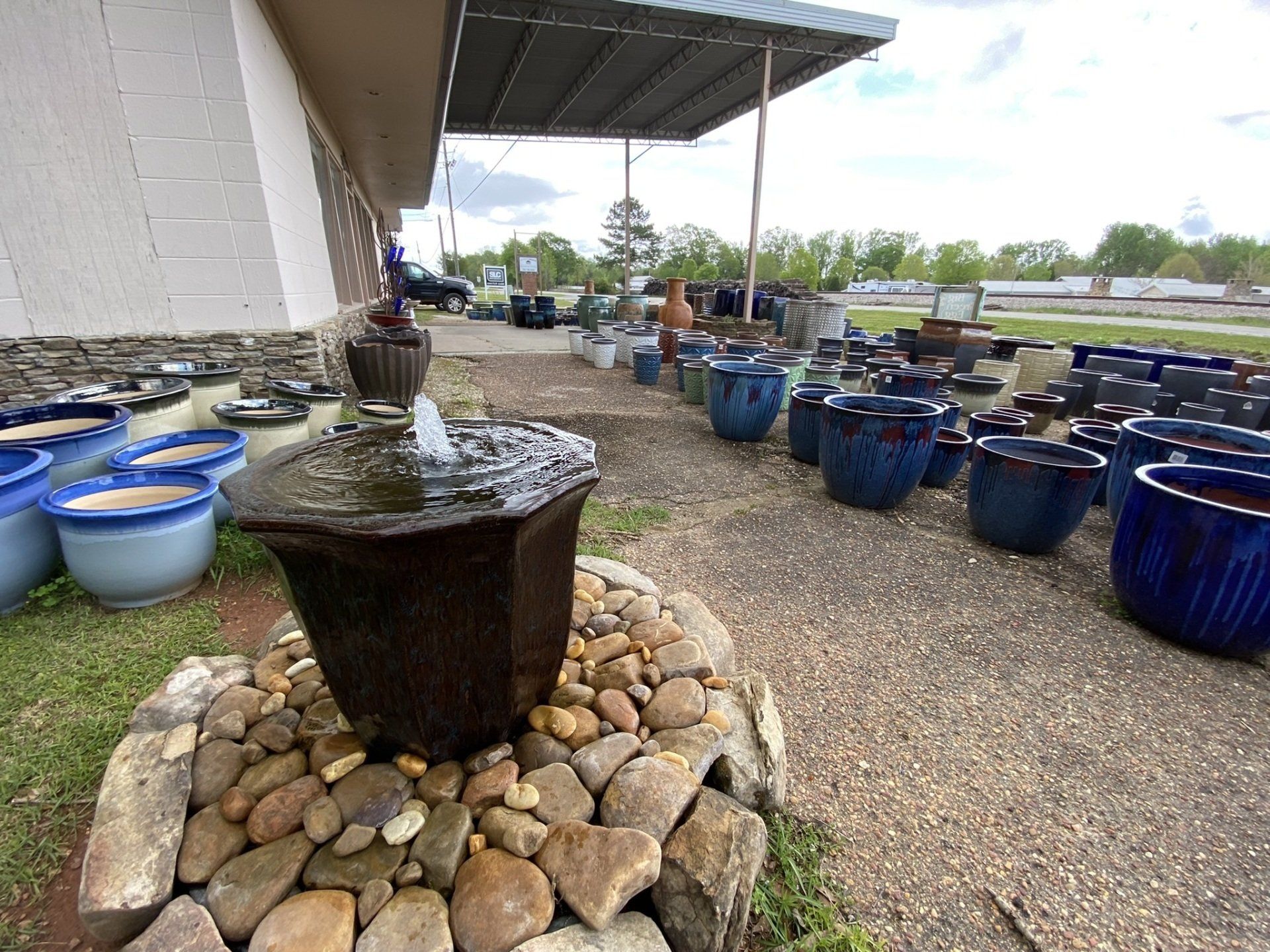 A fountain sprays water surrounded by stones.  Blue ceramic pots line a gravel path under a canopy.