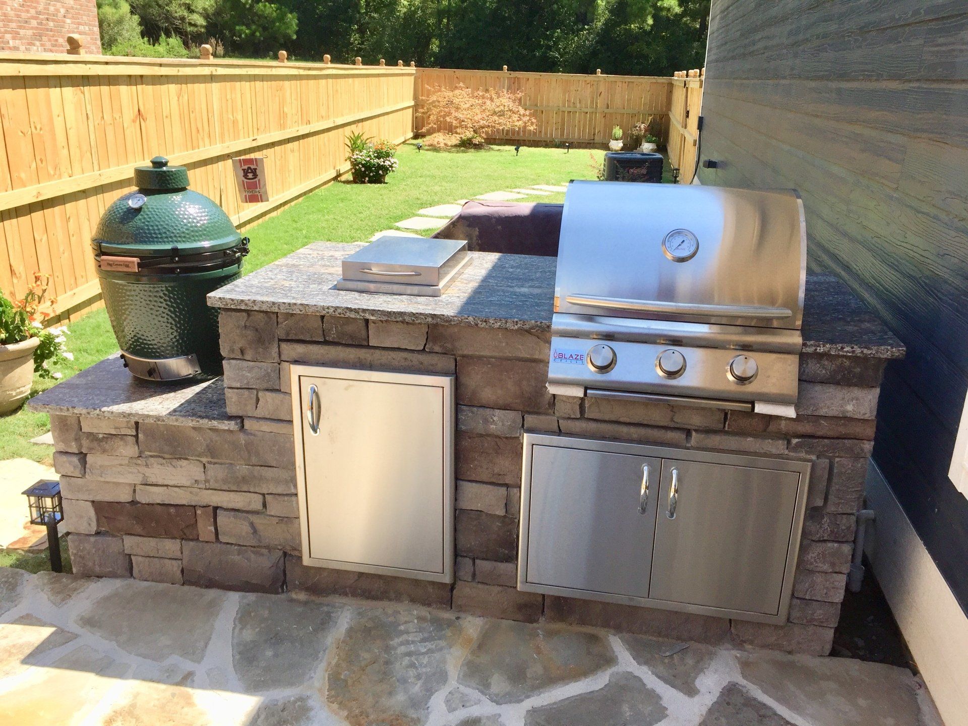A stainless steel grill is sitting on top of a stone counter in a backyard.