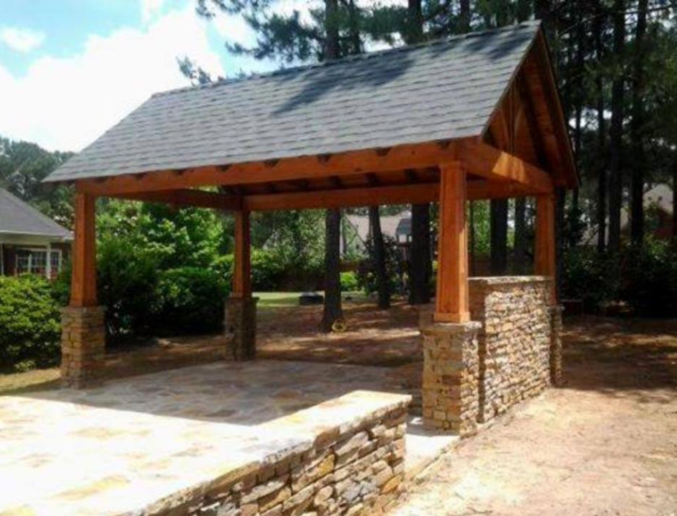 Wooden pavilion with stone columns and roof, set on a stone patio, with trees in the background.