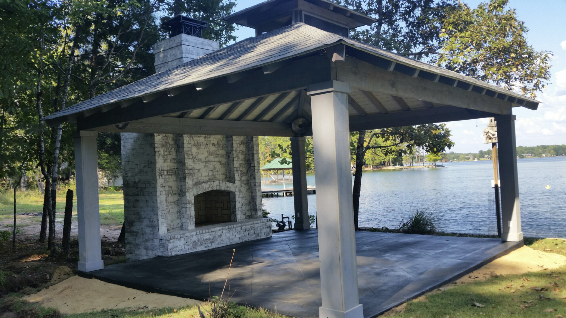 Outdoor pavilion with fireplace overlooking a lake. Gray columns, roof; green grass, trees in background.