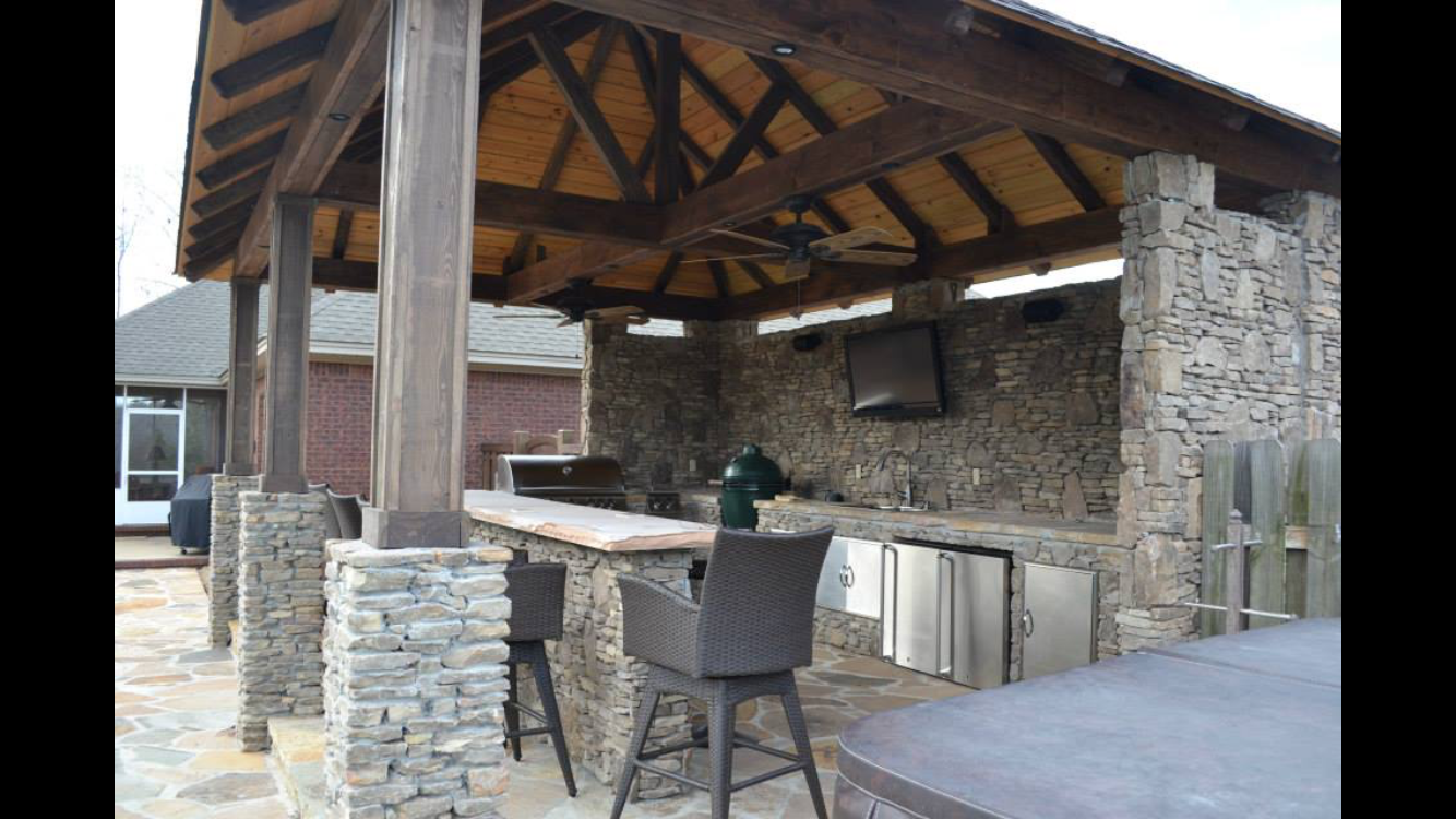 Outdoor kitchen with stone walls, wooden roof, bar, and built-in appliances, near a pool.