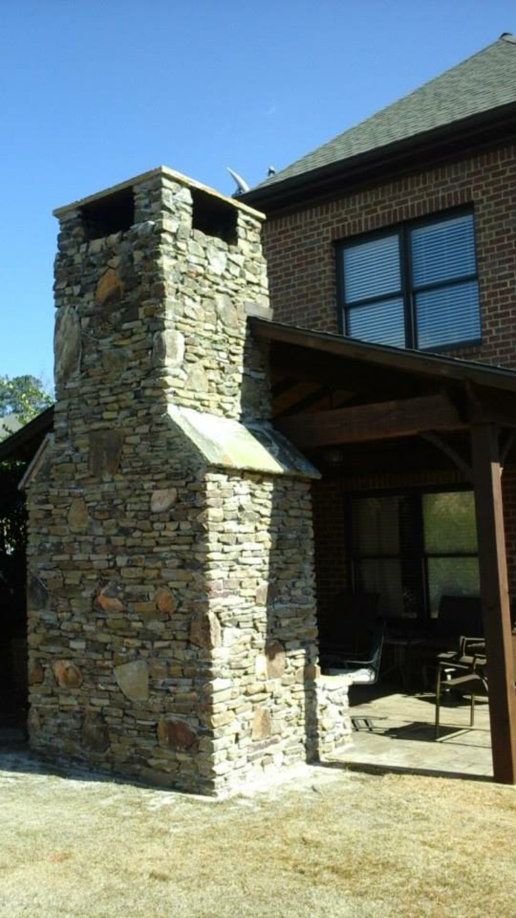 Stone chimney attached to a brick house, near a covered patio, on a sunny day.