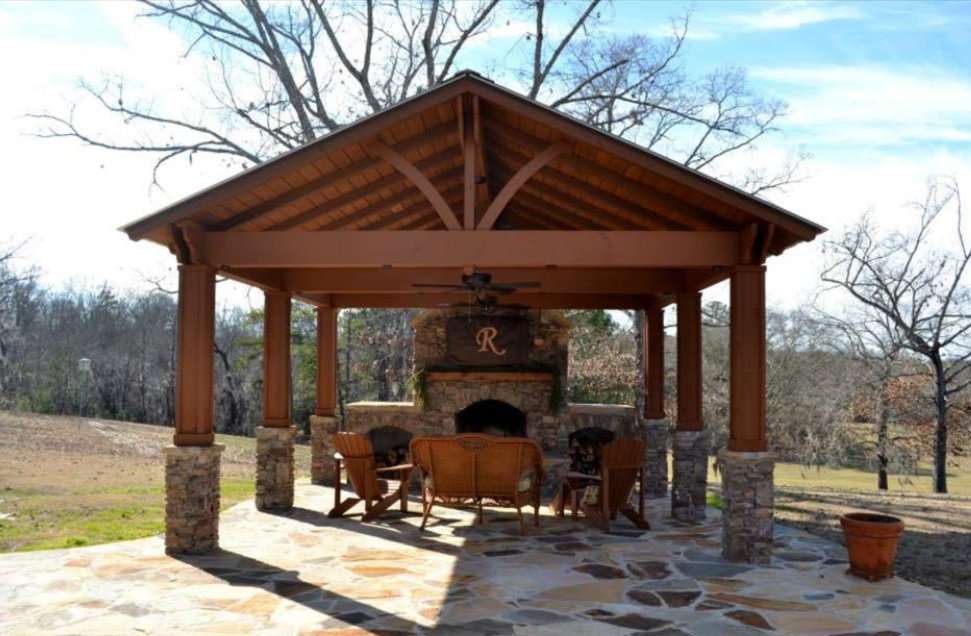 A brown wooden outdoor gazebo with a stone fireplace, seating, and a fan.