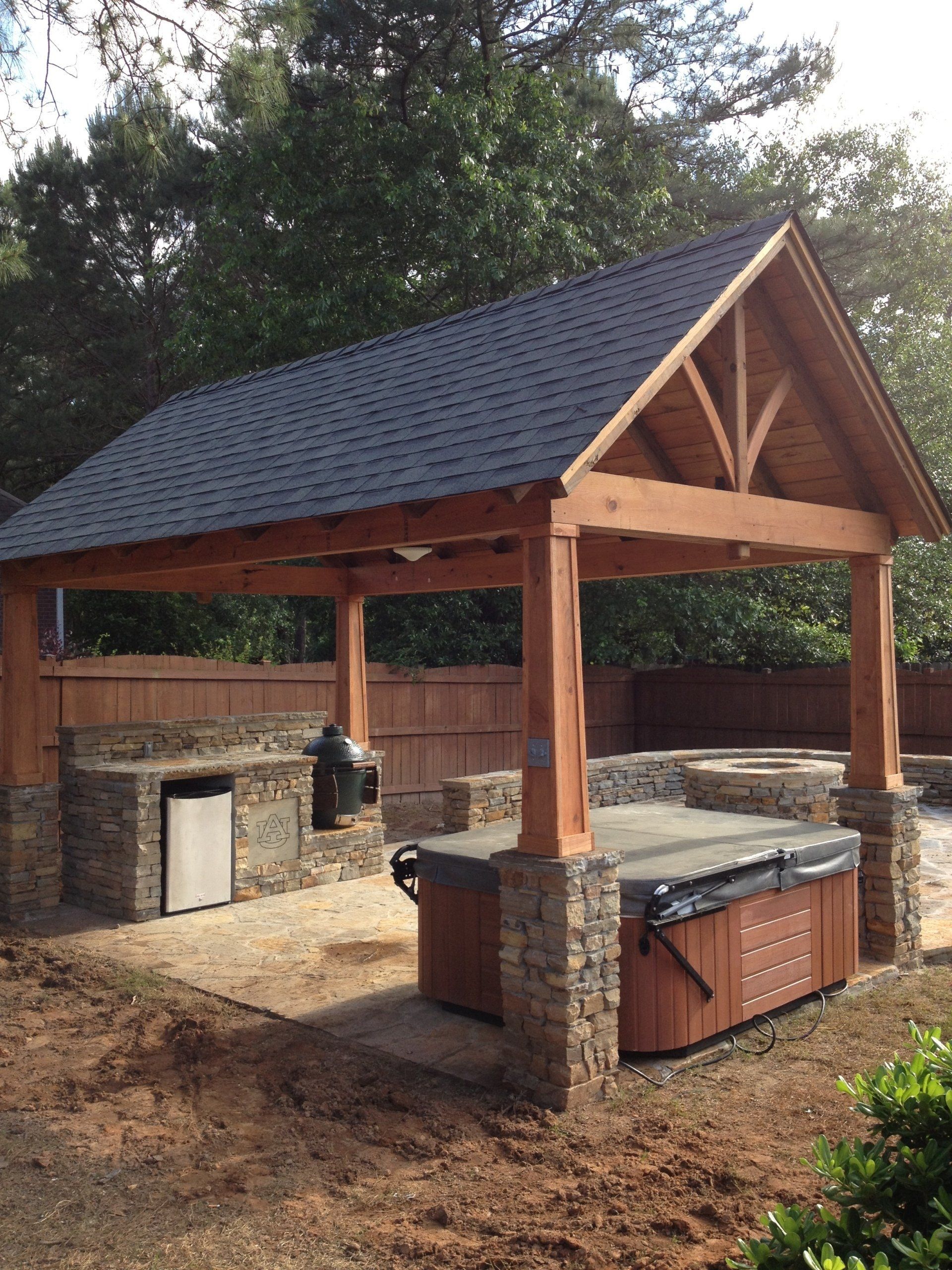 Outdoor cedar pergola with hot tub and grill station; stone accents, dark shingle roof, backyard setting.