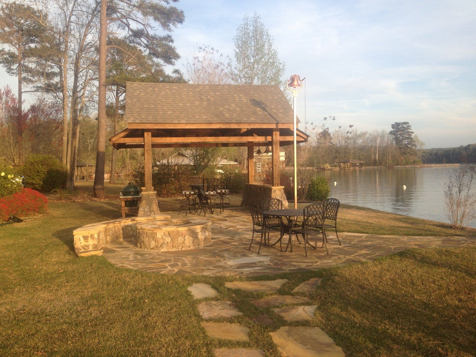 Gazebo with seating by lake, stone path, grassy area, trees, and a blue sky.