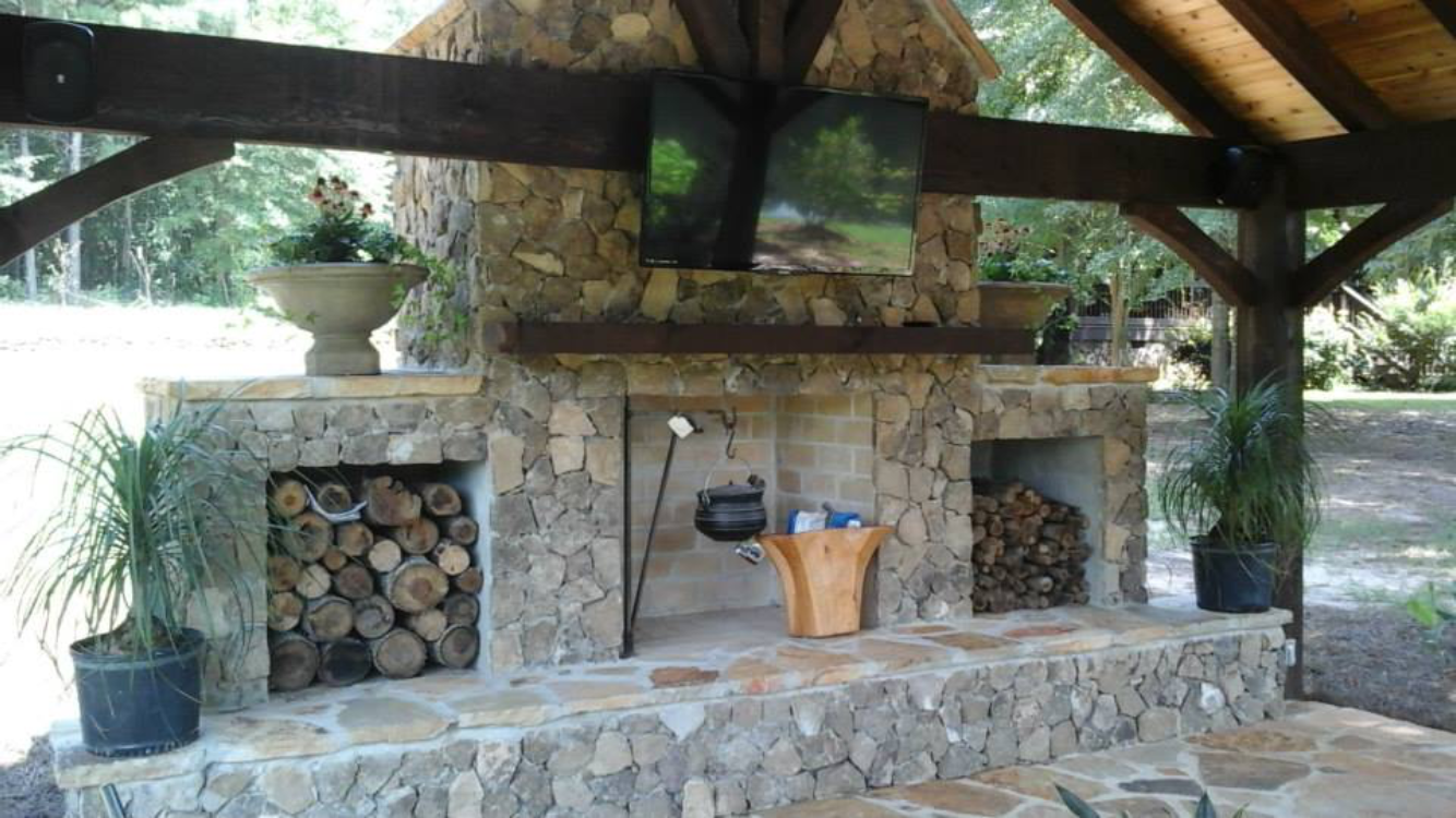 Outdoor stone fireplace with firewood storage, hanging pot, and TV screen under a wooden pergola.