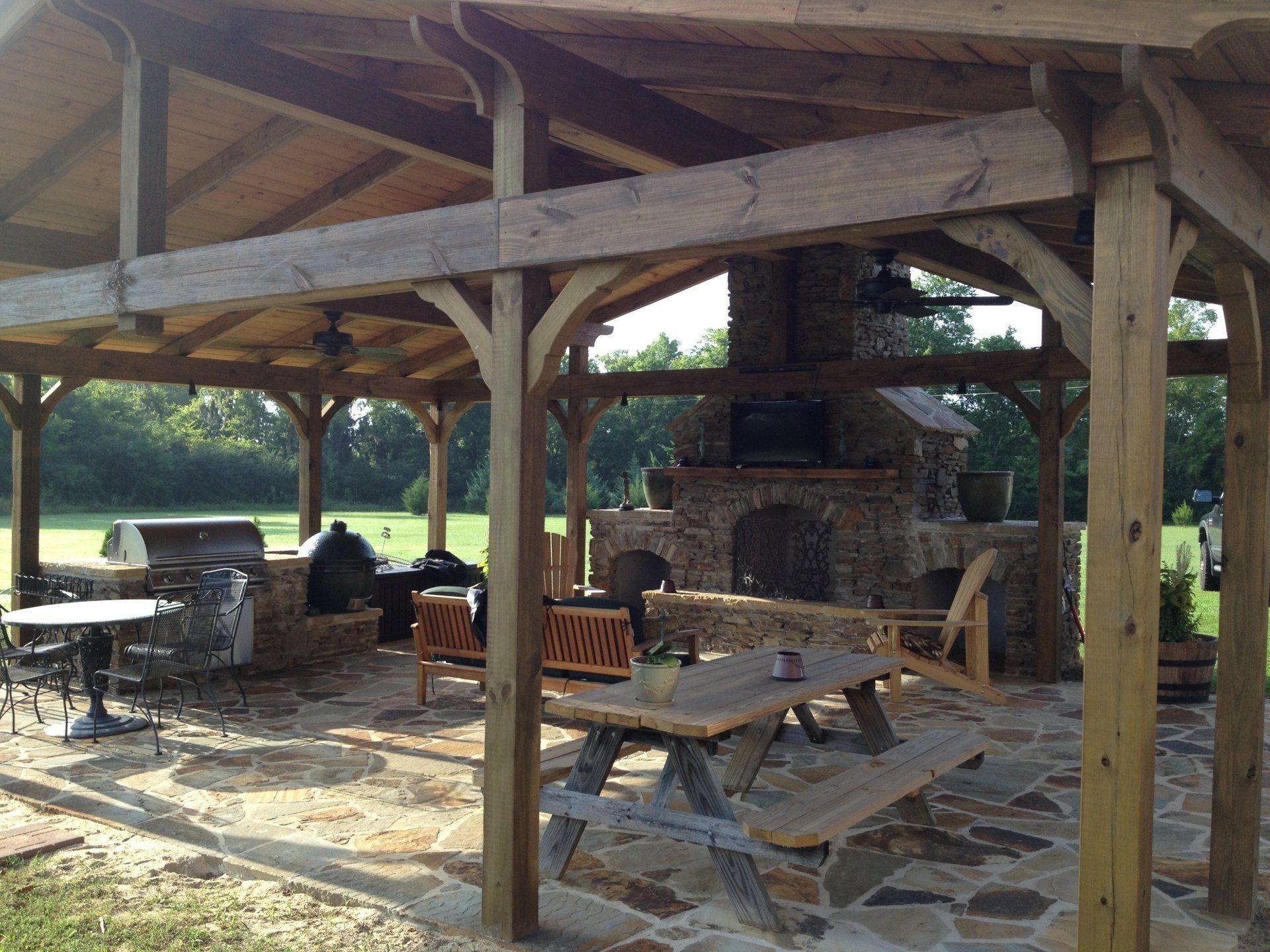 Outdoor kitchen with stone fireplace and grill under a wooden pergola; a picnic table is in front.