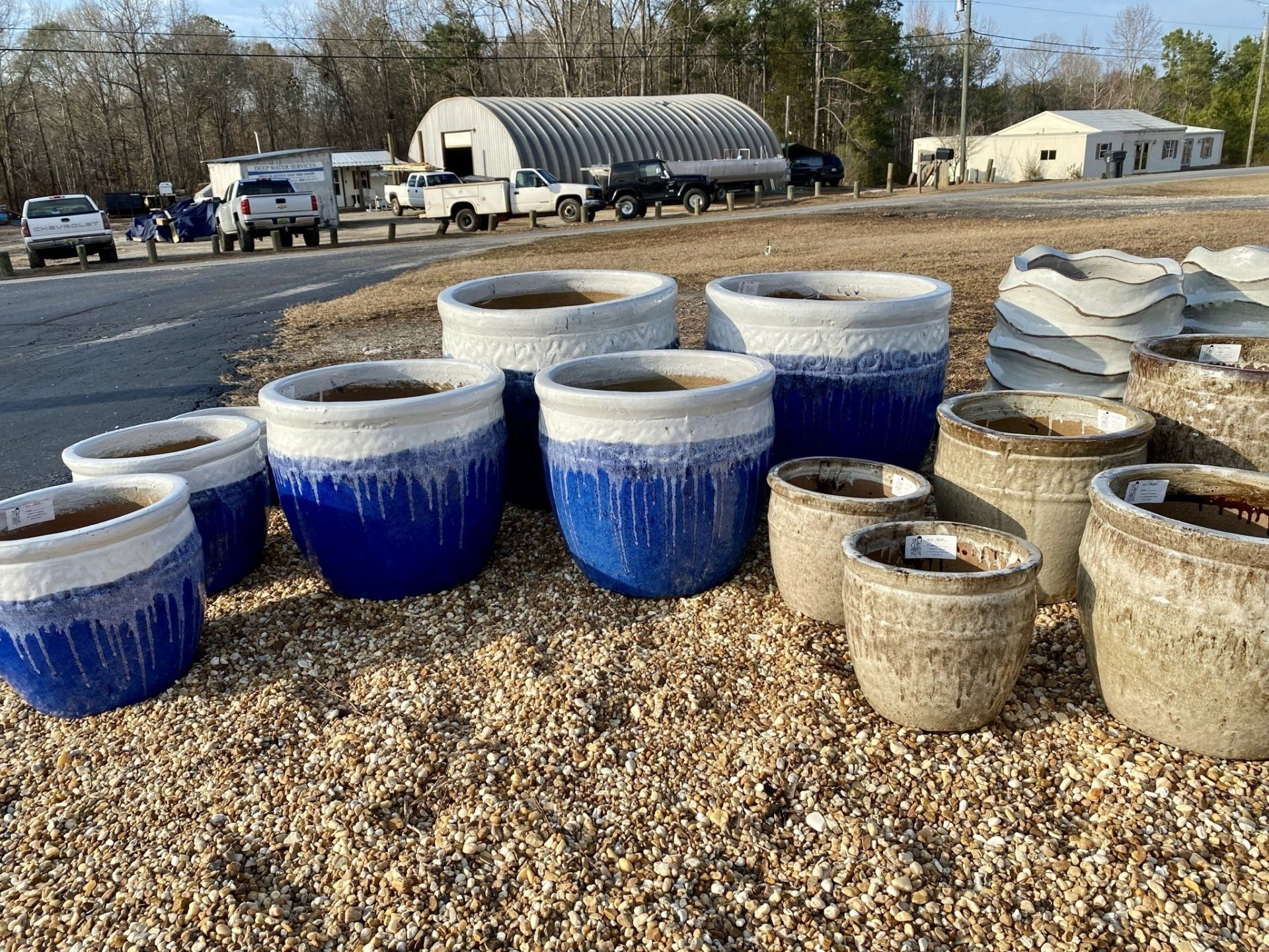 Row of blue and tan glazed ceramic planters on gravel, with buildings and vehicles in the background.
