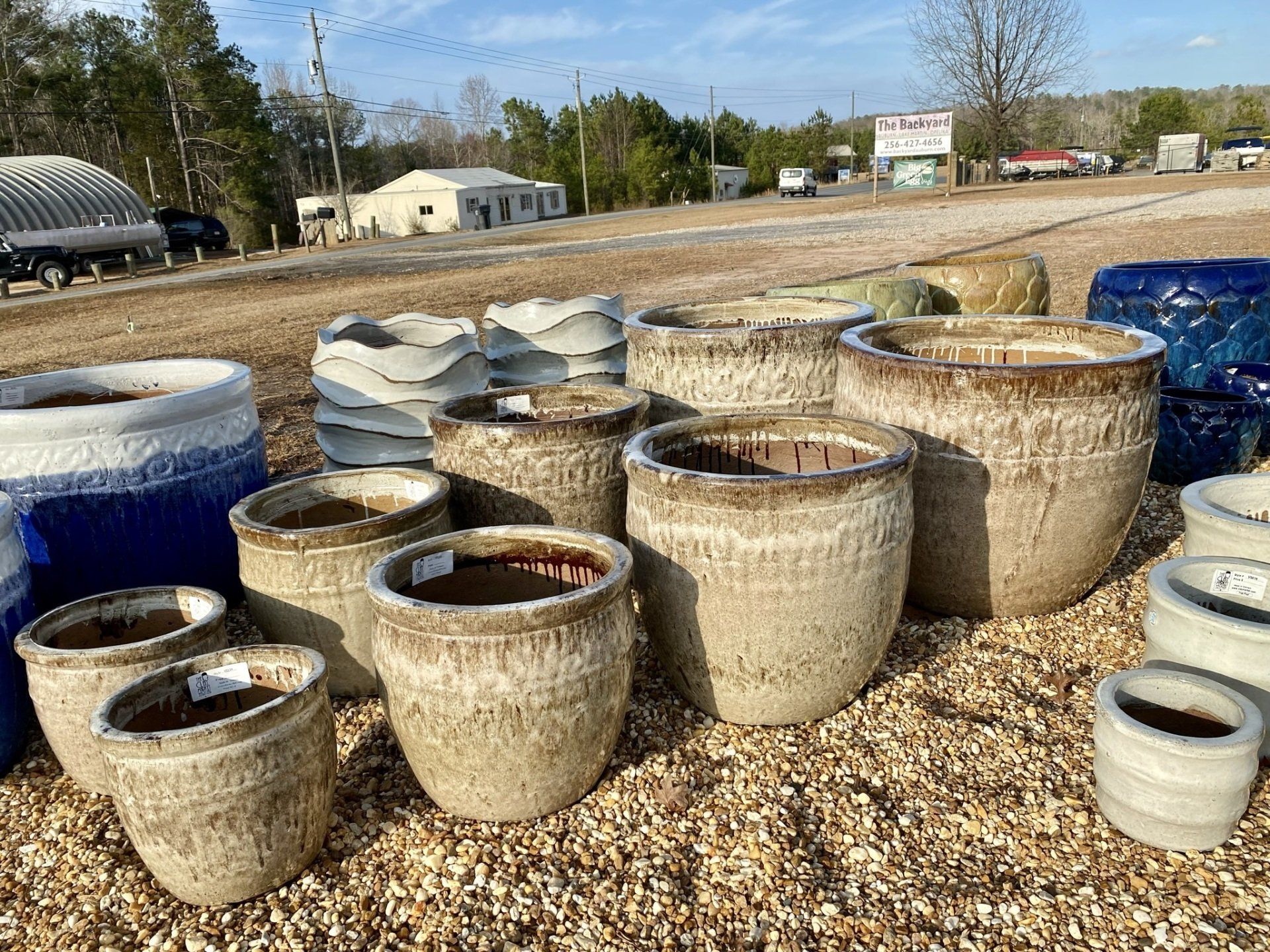 Group of ceramic pots in various sizes and colors on a gravel lot in front of buildings.
