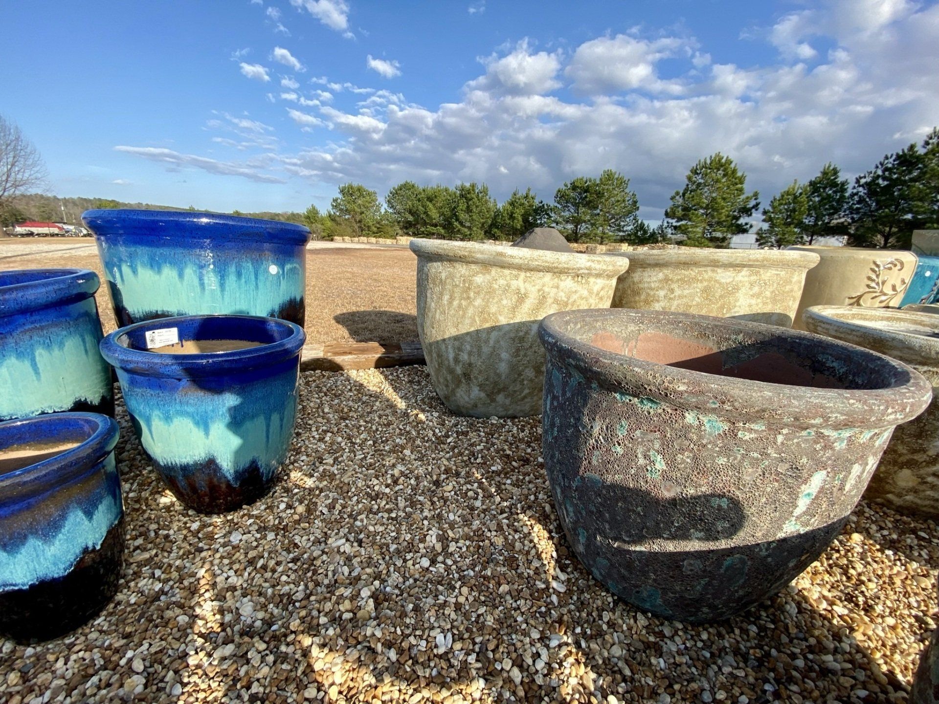 Various colorful ceramic planters on a gravel surface outdoors under a blue cloudy sky.