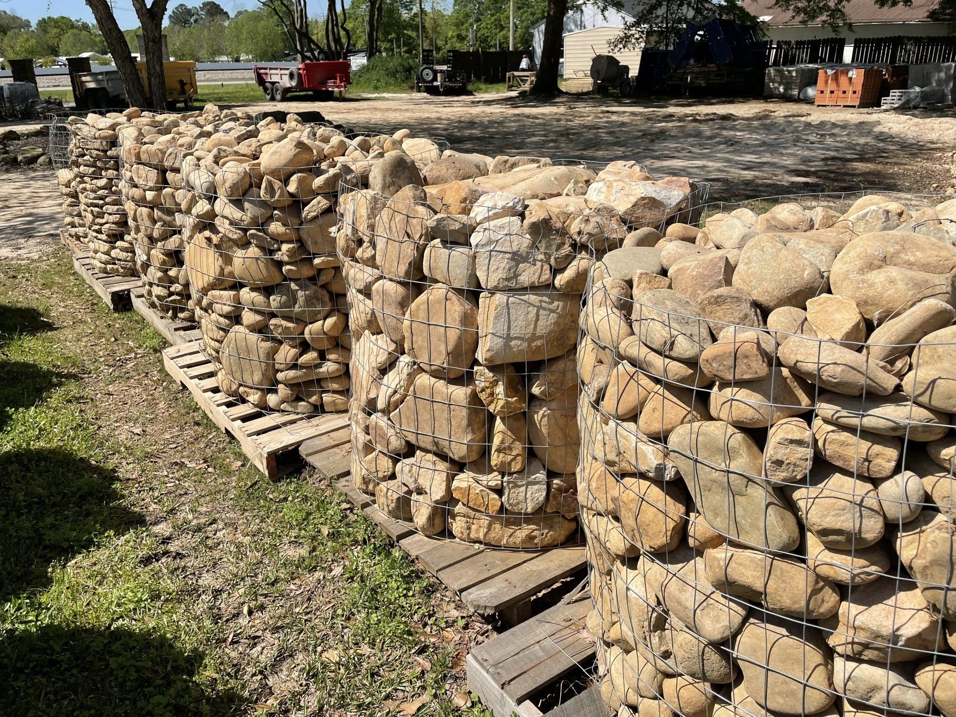 Cylindrical rock cages on pallets, filled with brown stones, line up outdoors on grass.