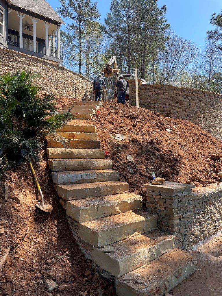 Stone steps being built on a hillside with construction workers and an excavator visible.