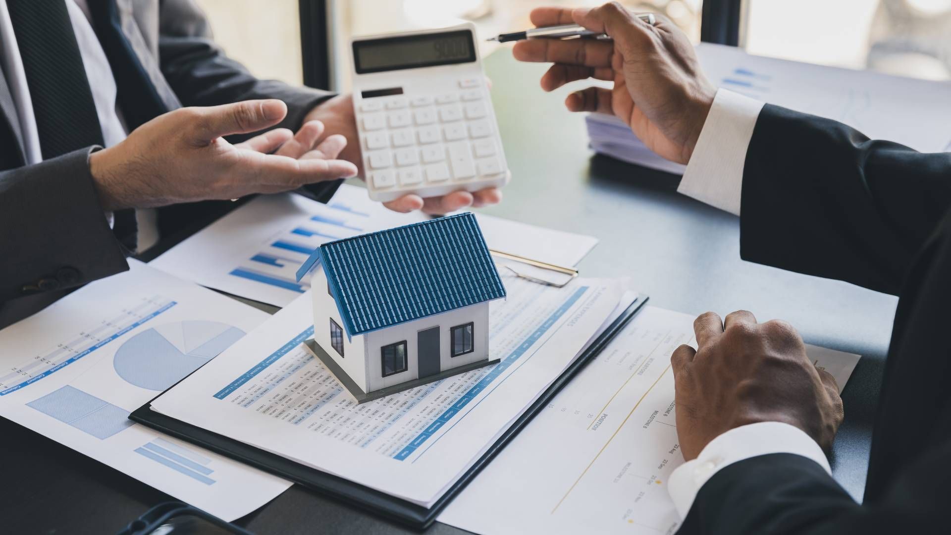 Two men are sitting at a table with a model house and a calculator.