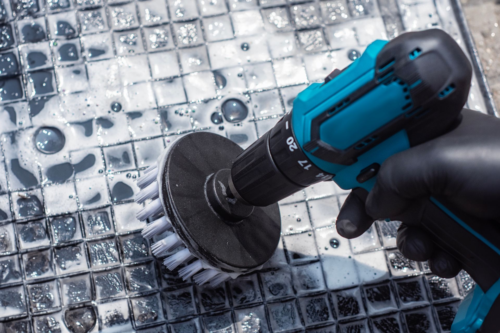 A gloved hand uses a power drill with a circular brush attachment to scrub a tiled surface covered in soapy foam.