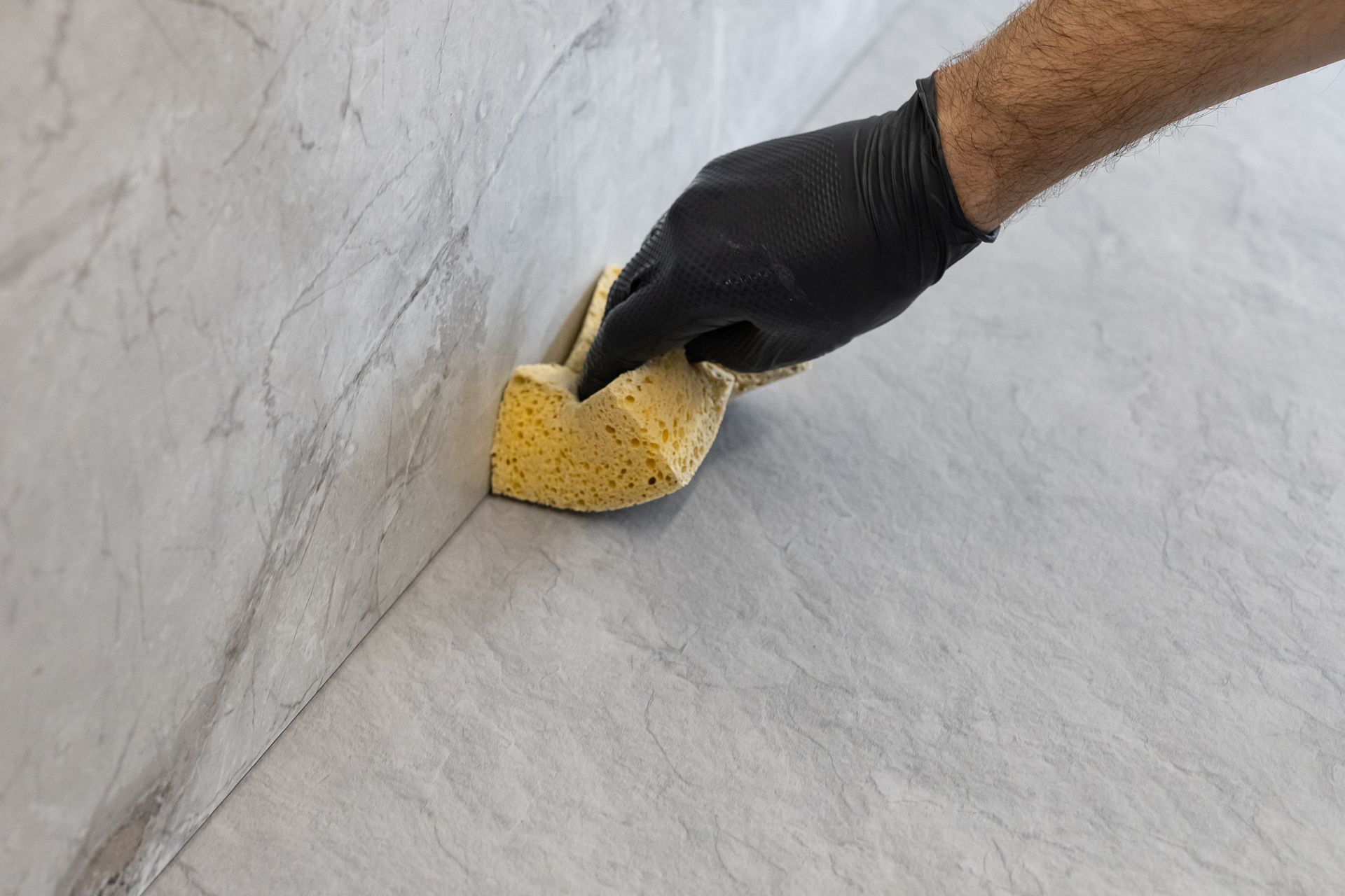 A hand wearing a black glove uses a yellow sponge to smooth gray tile grout along a wall corner.