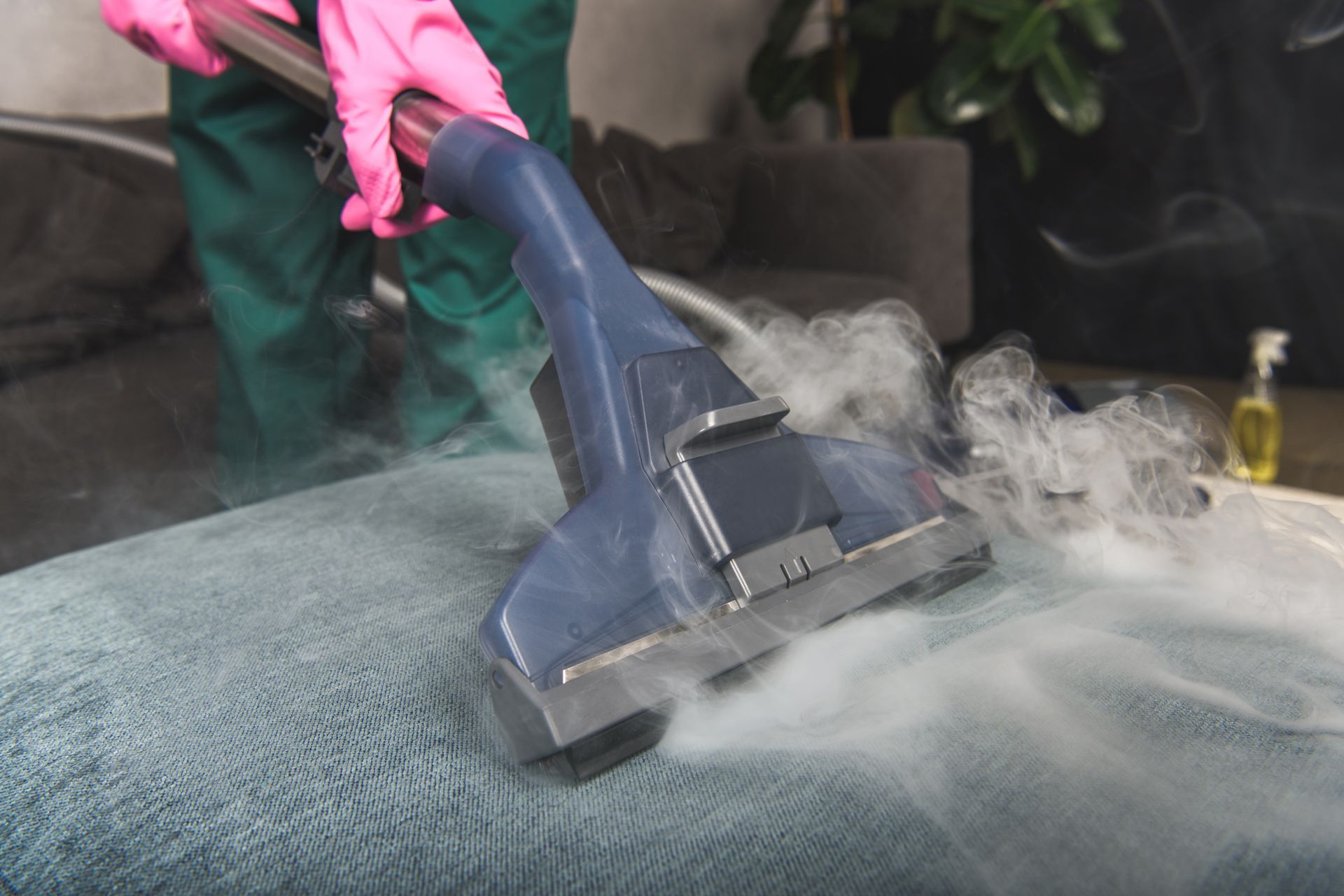 A person wearing pink gloves uses a steam cleaner to sanitize a blue upholstered surface.