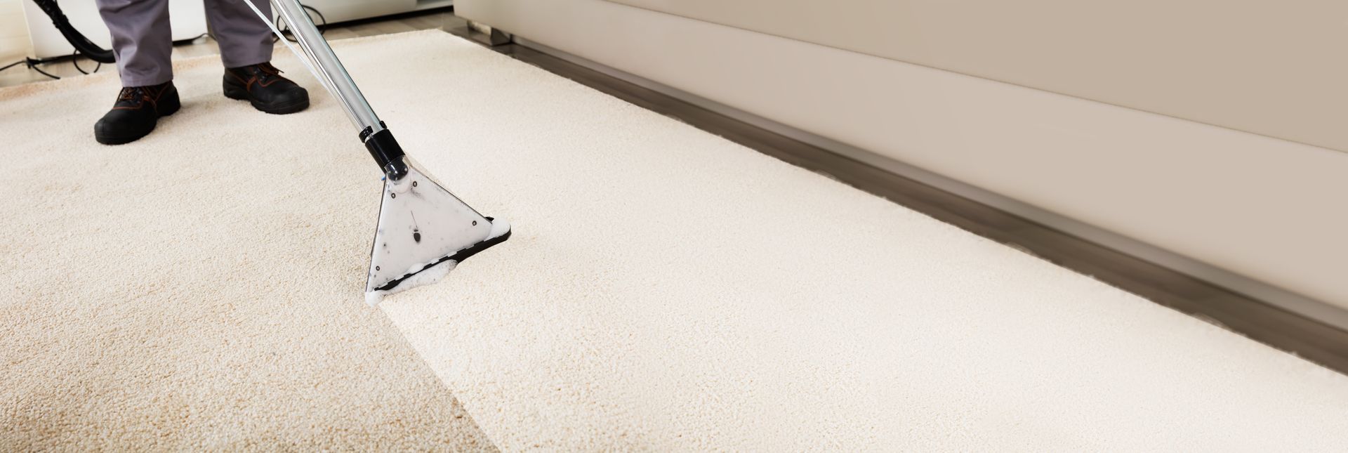 A person uses a commercial steam cleaning wand to deep clean a light-colored carpet along a wall baseboard.