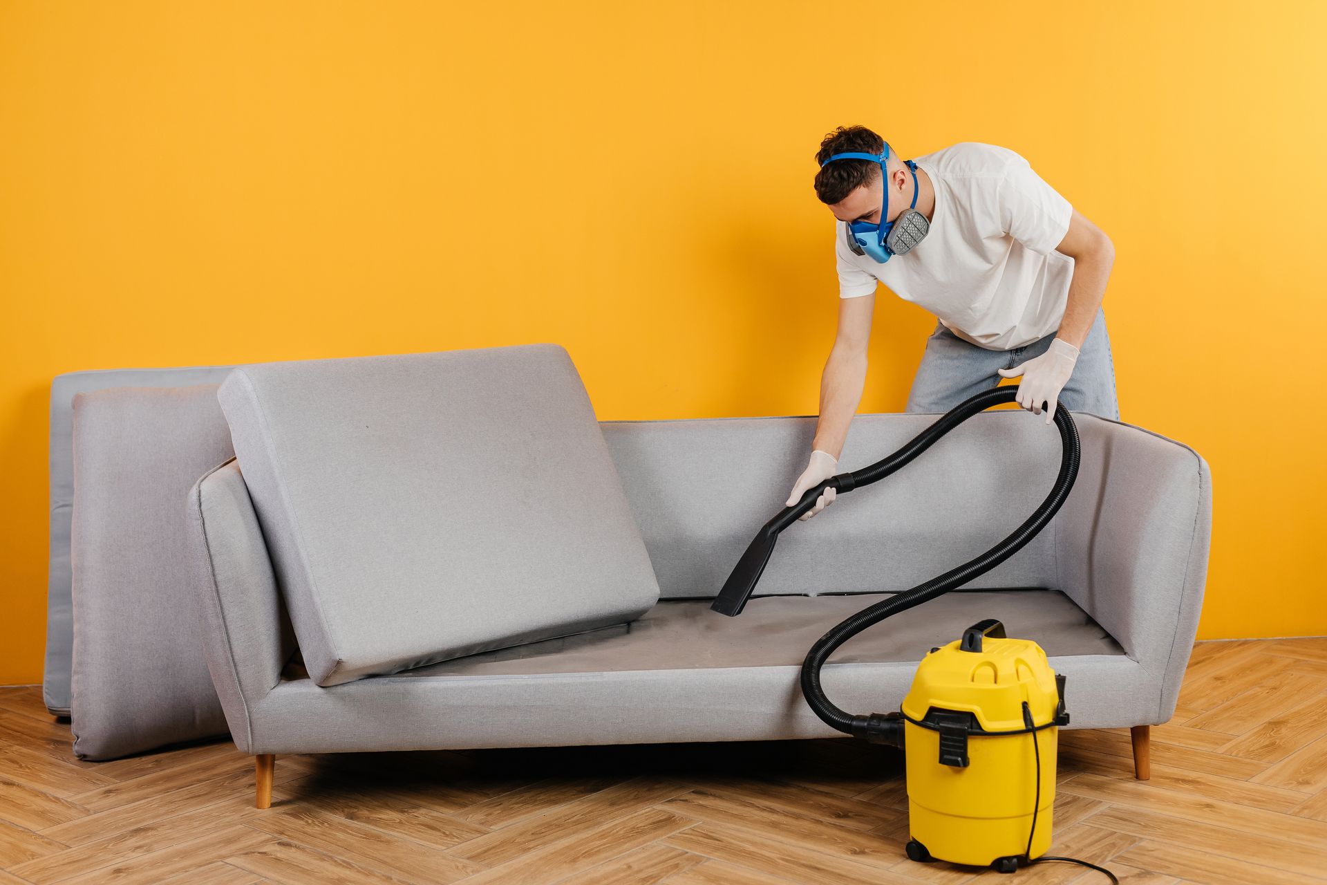 A person wearing a protective mask and gloves uses a yellow vacuum to clean a grey sofa against a bright yellow wall.