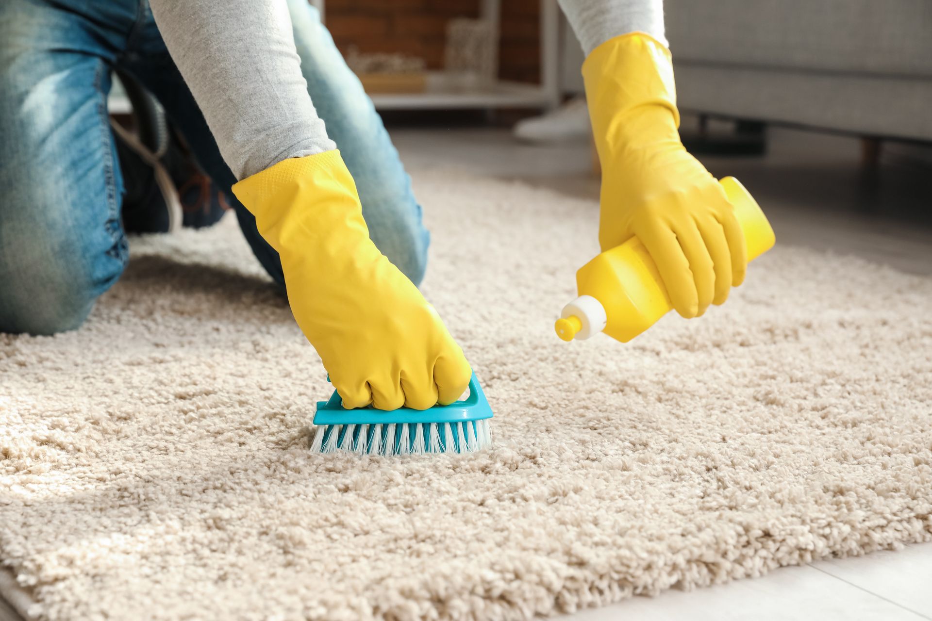 A person wearing yellow rubber gloves kneeling on a beige carpet, scrubbing a stain with a blue brush and yellow cleaner.