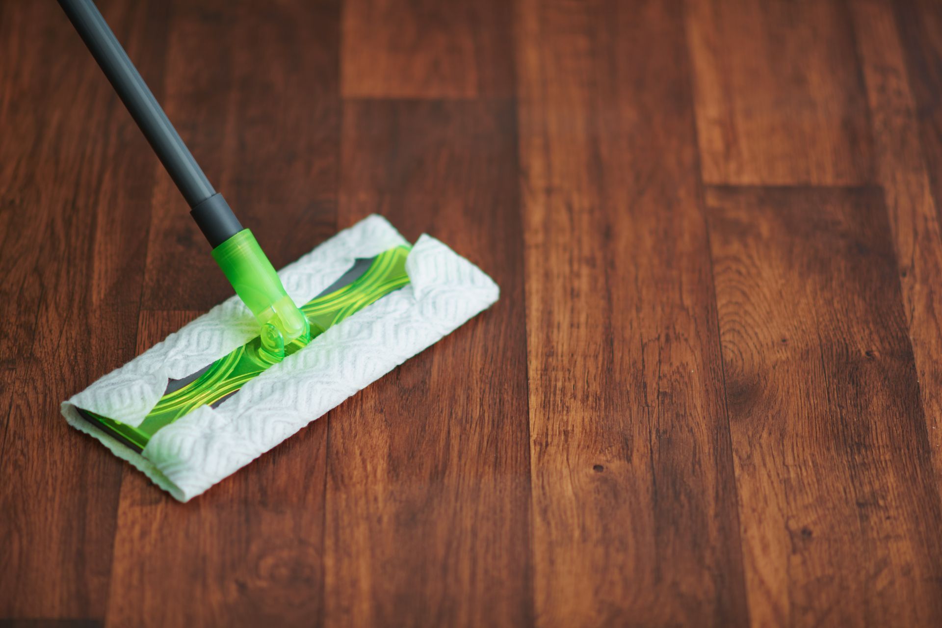 A green and grey mop head with a white paper towel attached cleaning a dark wood floor.