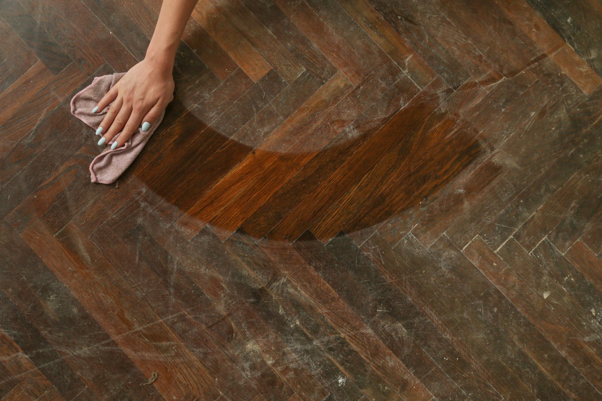 A person wipes a cloth across a wooden parquet floor, revealing a clean, polished section against a duller, dusty surface.