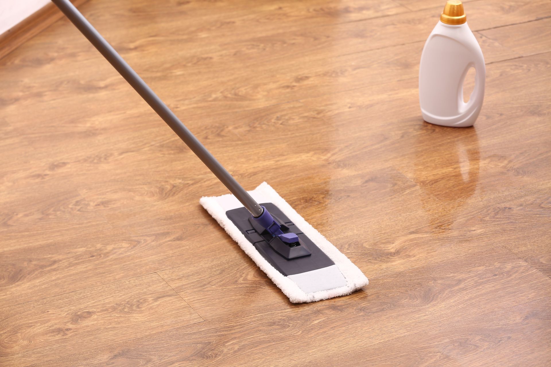 A flat mop cleans a shiny, wood-look floor, with a white cleaning product bottle nearby.