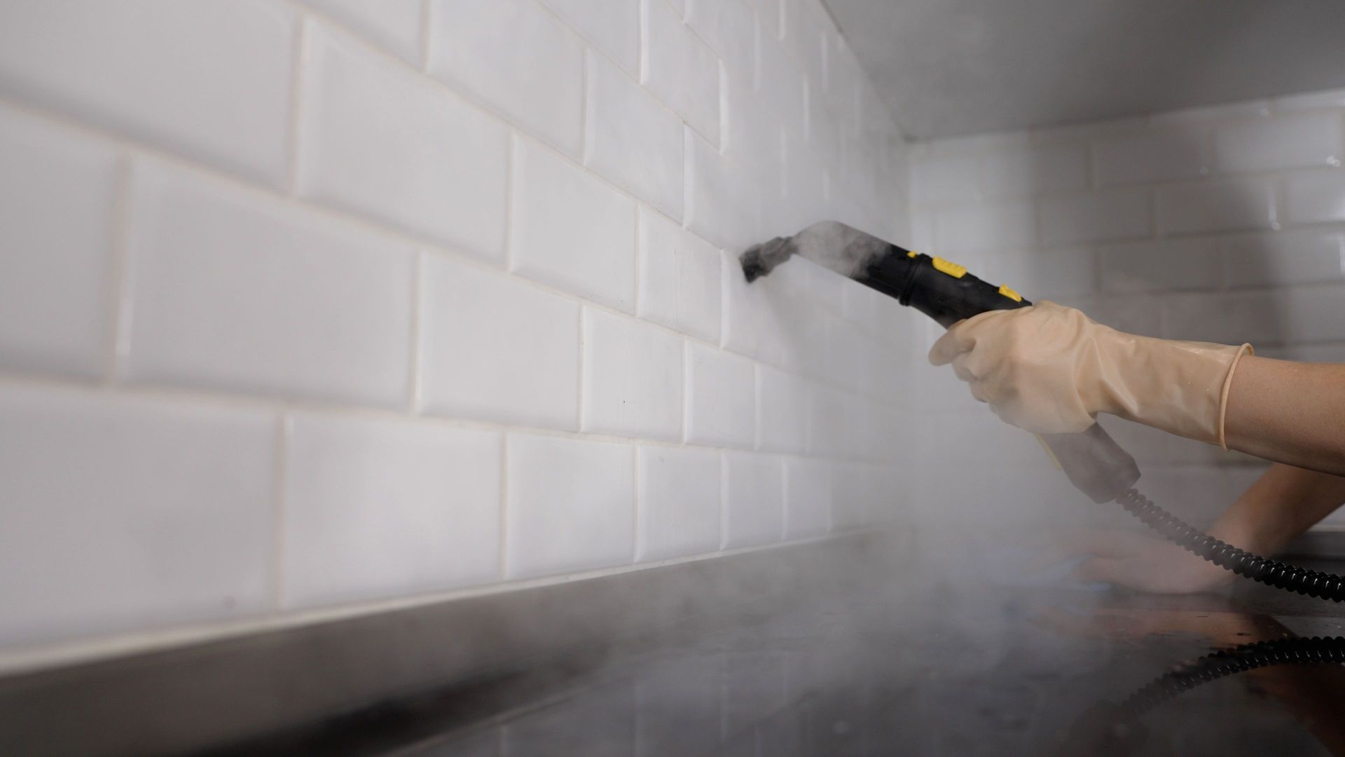 A gloved hand uses a steam cleaner to sanitize white subway-style kitchen tiles.