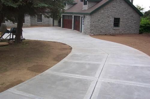 Concrete driveway curves toward a stone house with garage doors. Green grass and brown gravel surround.