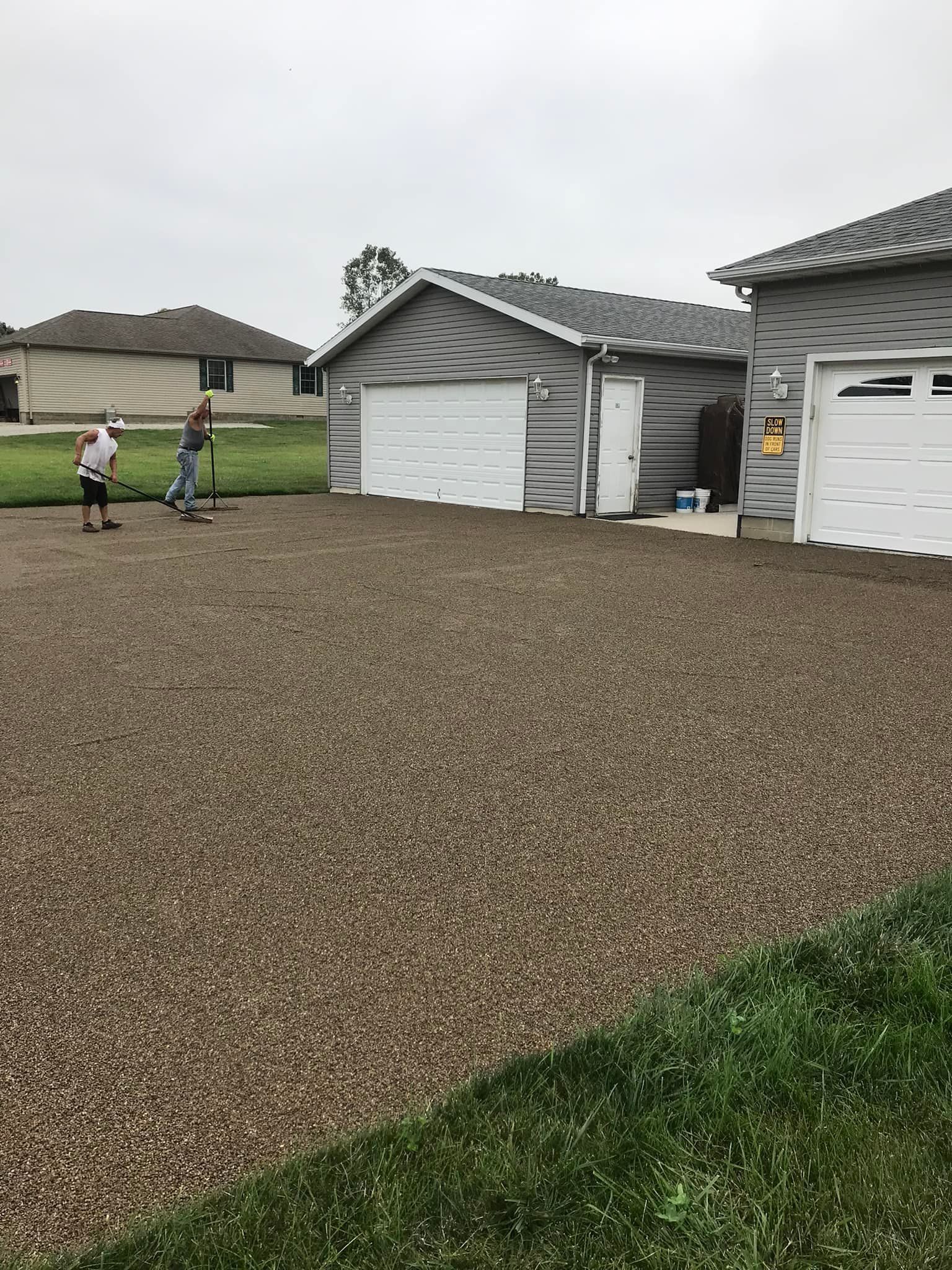 A gravel driveway in front of a garage. Two people are working on the gravel. Houses in the background.