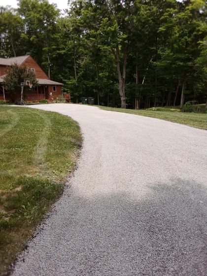 Gravel driveway curves toward a wooden house, bordered by green grass and trees.