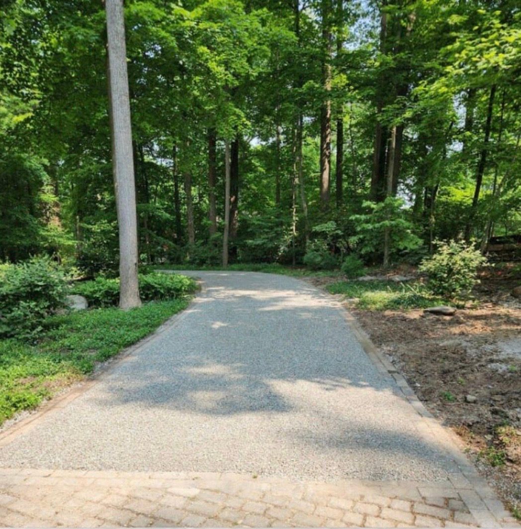 Gravel driveway leading into a wooded area lined with trees and shrubs on a sunny day.