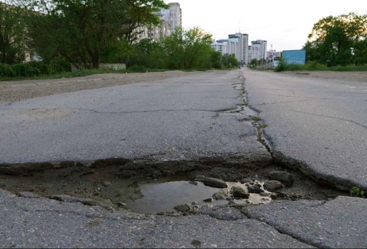 Damaged asphalt road with large pothole filled with water; city buildings in the background.
