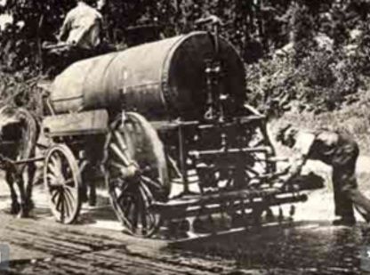 Horse-drawn water wagon spraying a road, two workers, outdoors.