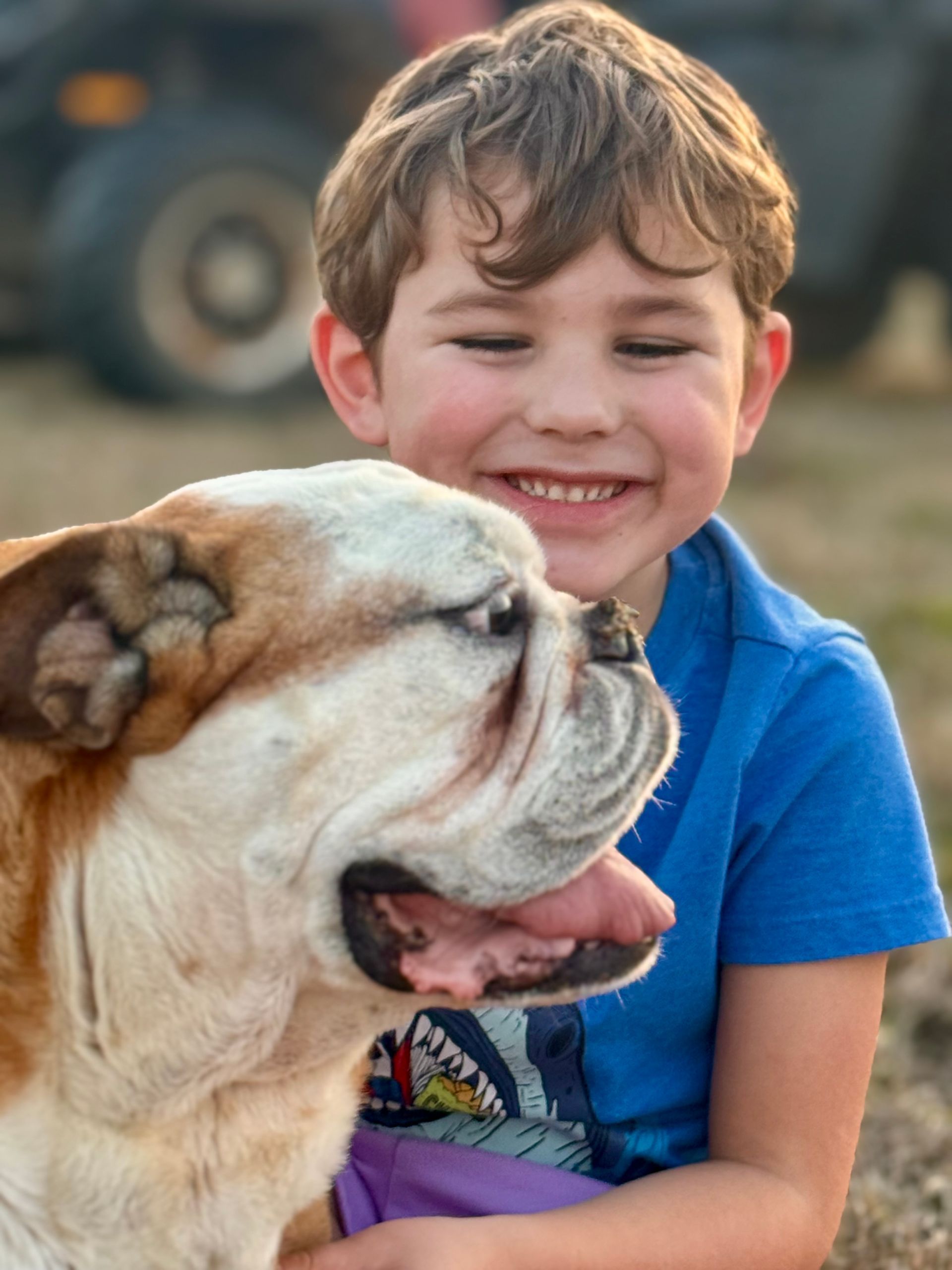 Boy smiles while hugging bulldog outside.