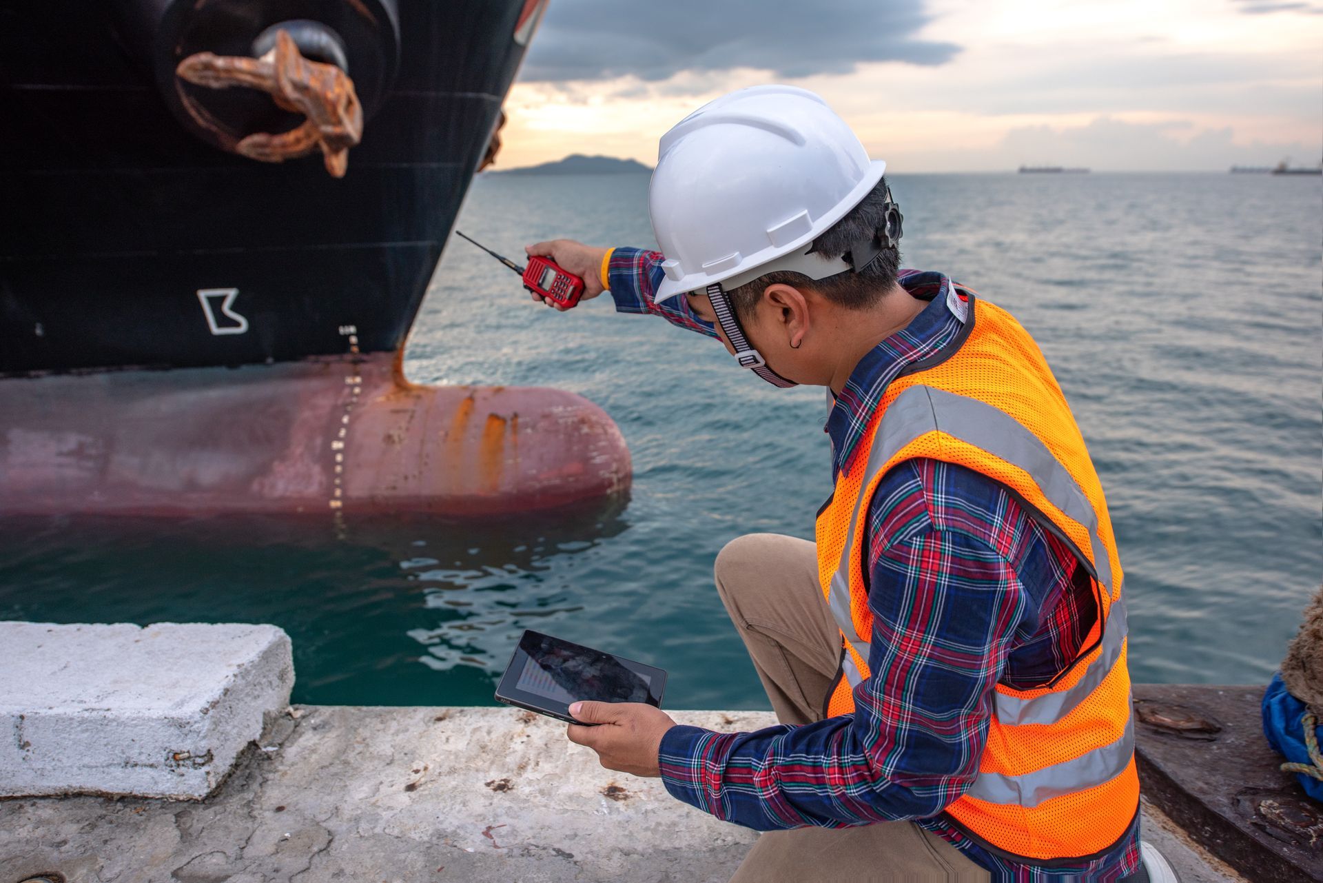 Person in safety gear inspecting a ship's hull at a dock, holding a device and looking at it.
