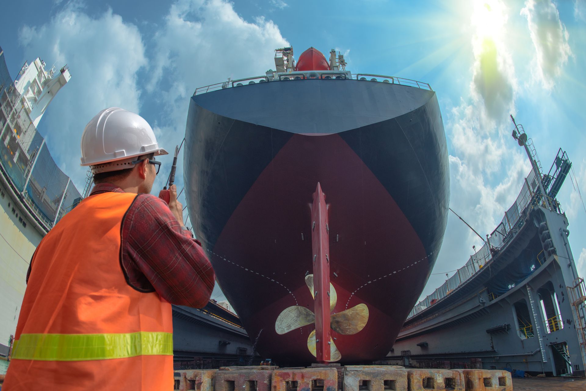 Man in hard hat and vest looks at large ship in drydock, sunny day.