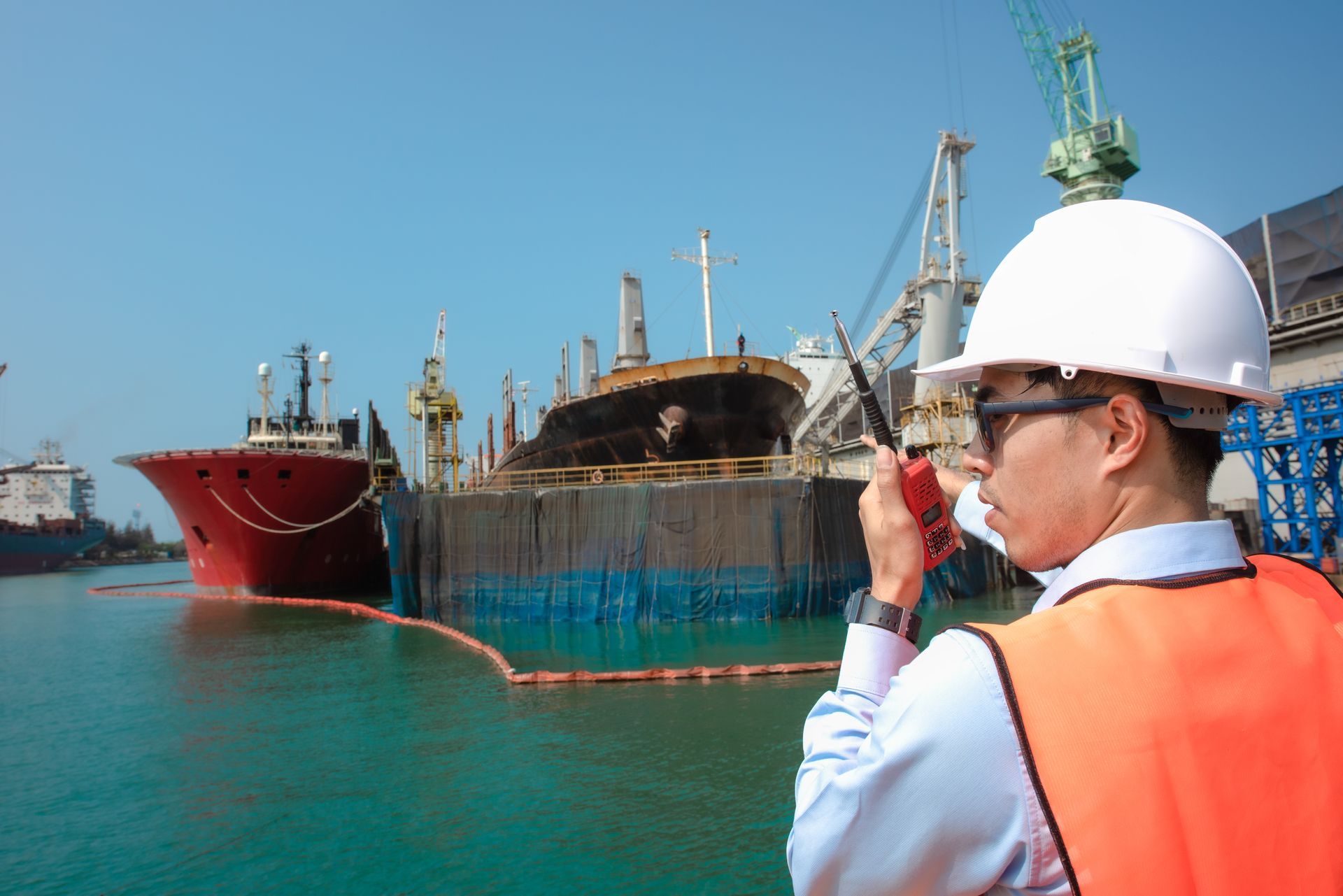 Harbor worker using a radio, wearing a hard hat and orange vest, with ships in the background.