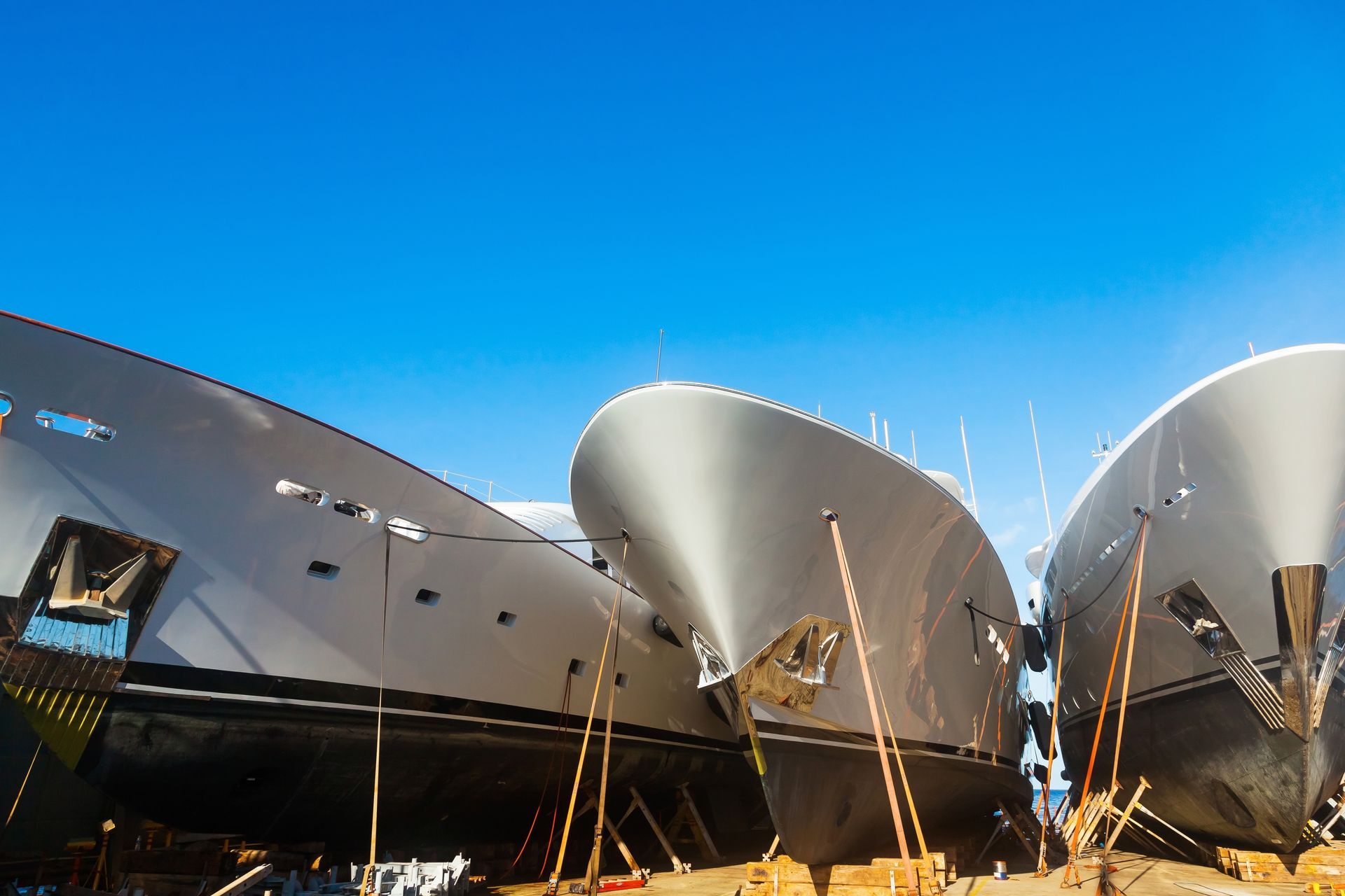 Three large gray boat hulls on wooden supports under a bright blue sky.