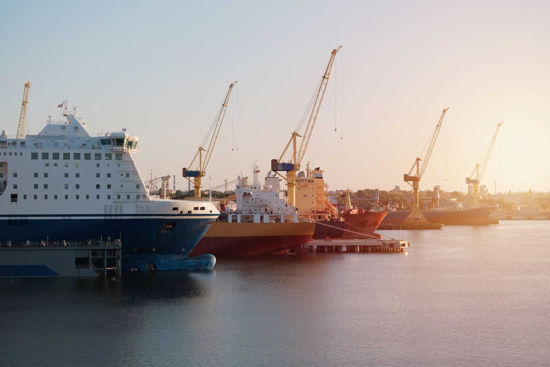 Ships docked at a harbor, with cranes in the background, lit by the setting sun.